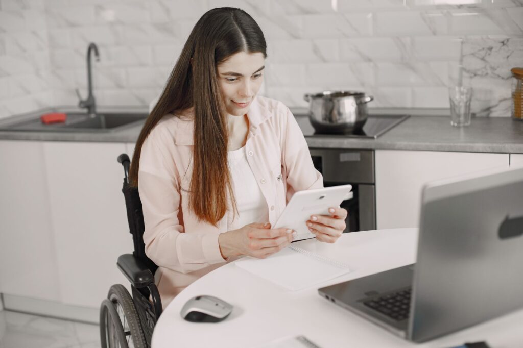 A woman in a wheelchair works on a tablet at the kitchen table